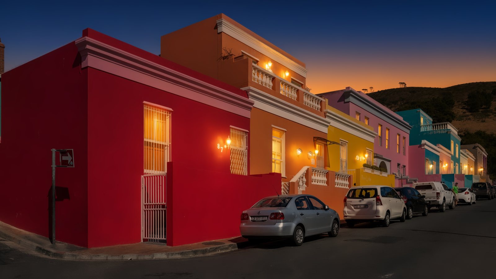 A row of brightly painted houses photographed in daylight, then relit as a twilight photo with a deep blue sky and warm light glowing in the windows, using AI — after