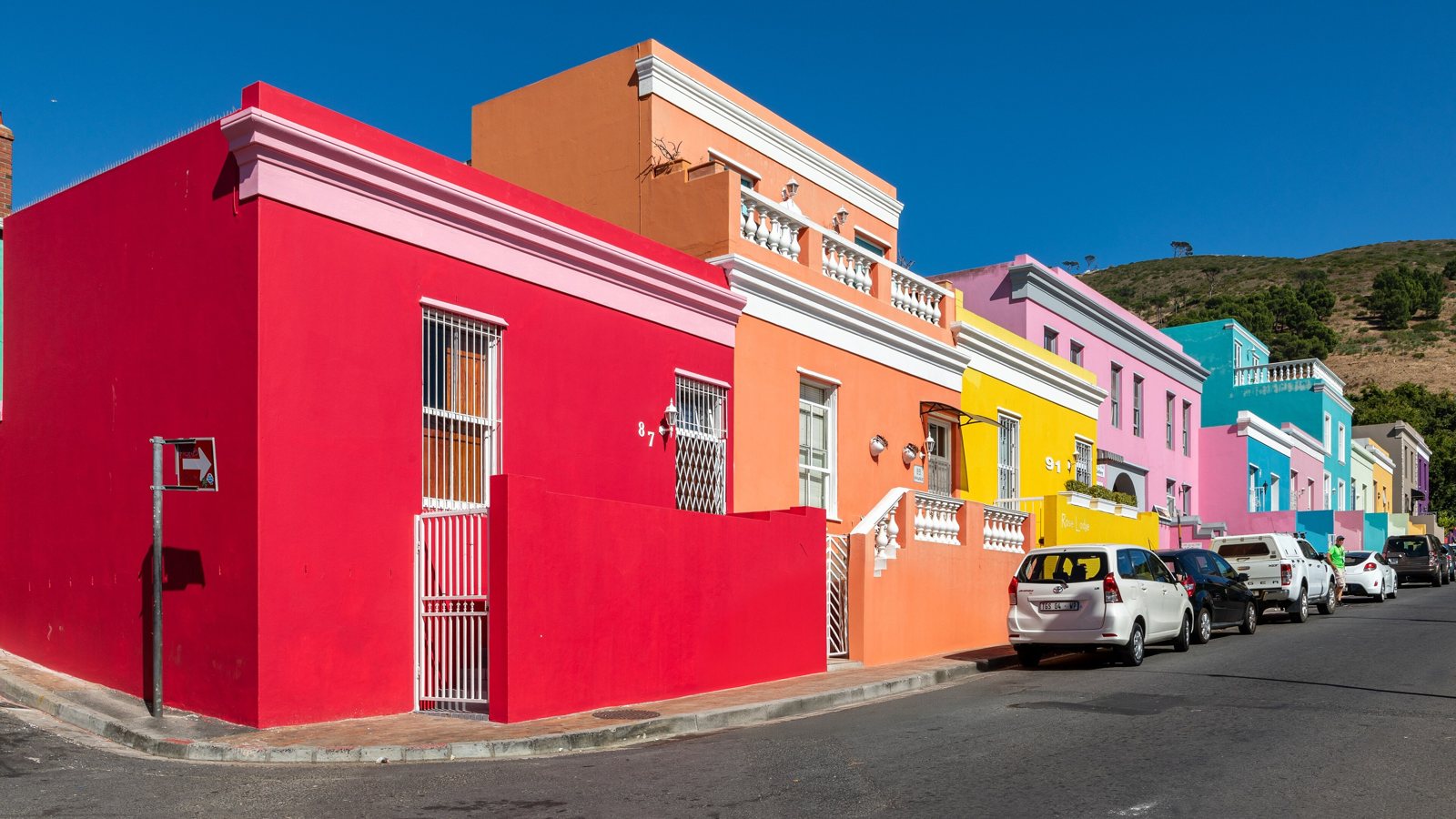A row of brightly painted houses on a street before and after removing the silver sedan parked at the curb with AI, the red wall and kerb filled in and the other cars untouched — after