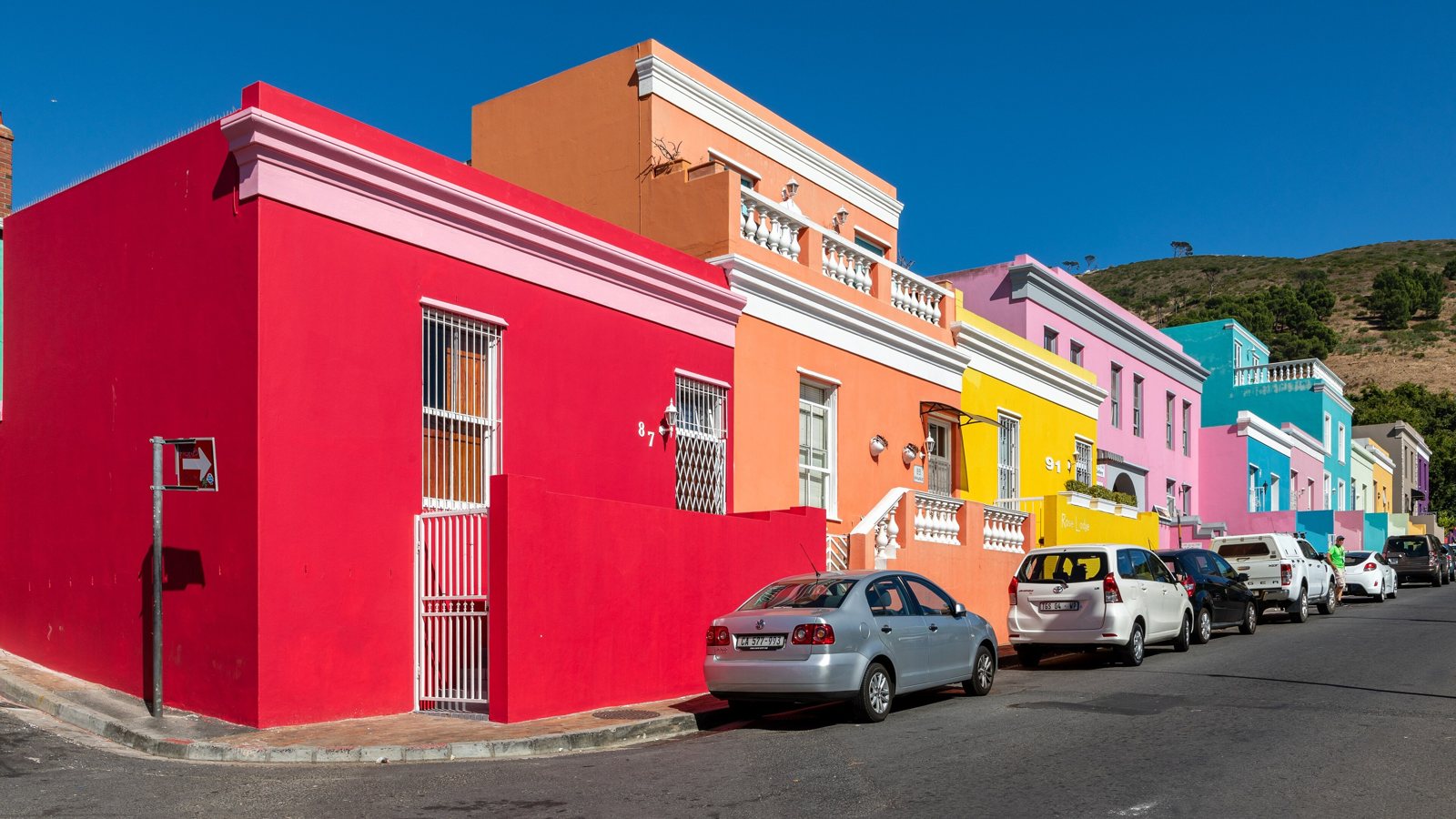 A row of brightly painted houses on a street before and after removing the silver sedan parked at the curb with AI, the red wall and kerb filled in and the other cars untouched — before