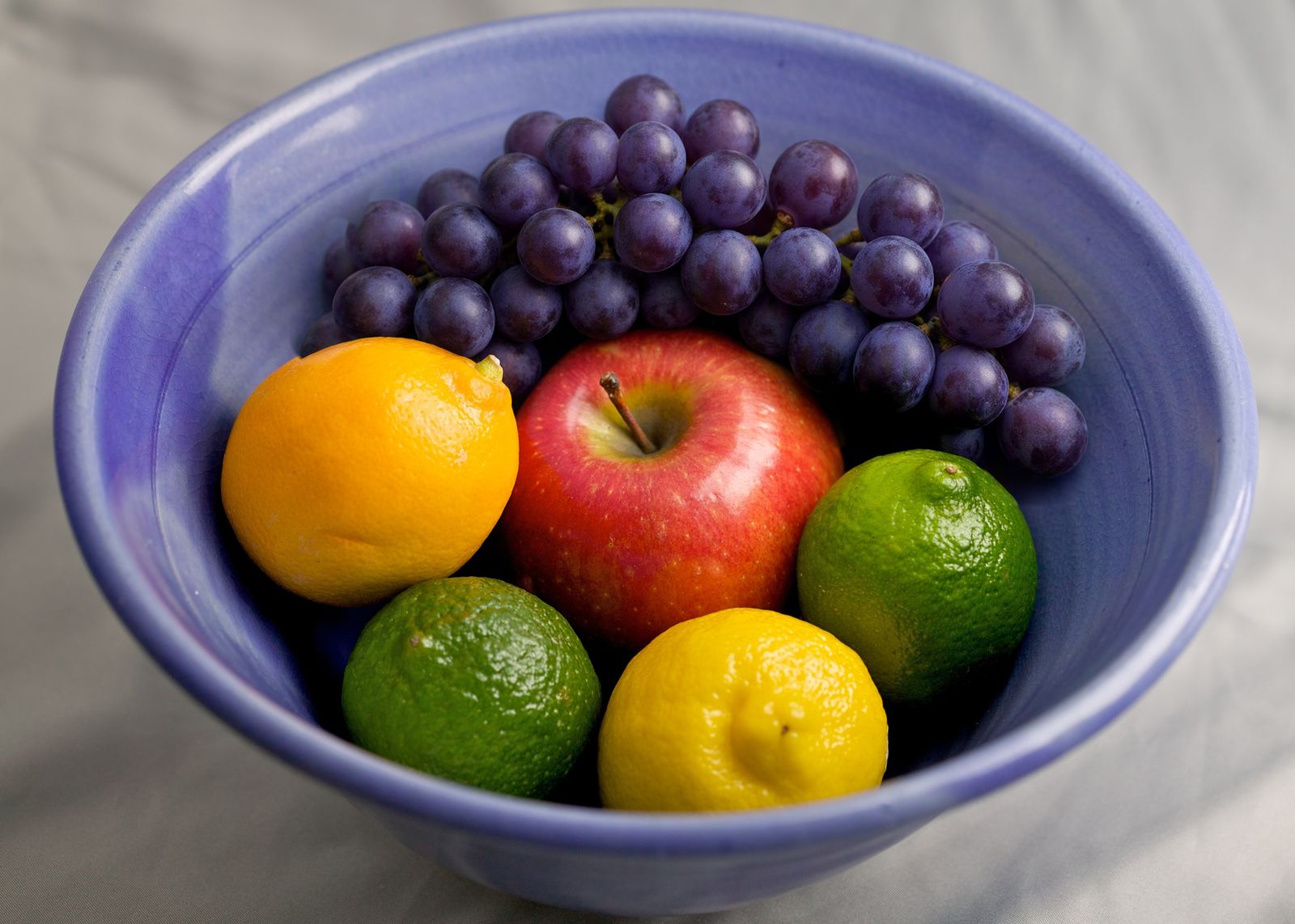 A bowl of fruit before and after replacing the banana with a bunch of purple grapes using an AI photo editor, the apple and citrus fruit unchanged — after
