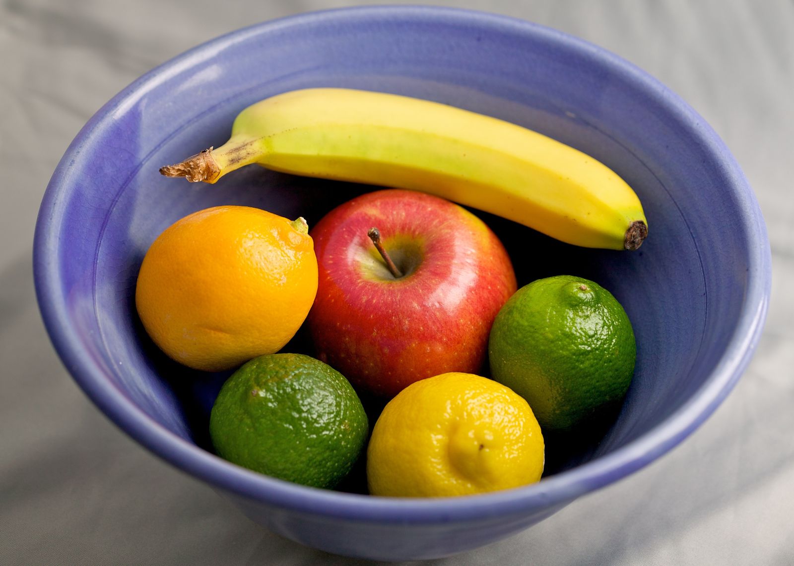 A bowl of fruit before and after replacing the banana with a bunch of purple grapes using an AI photo editor, the apple and citrus fruit unchanged — before