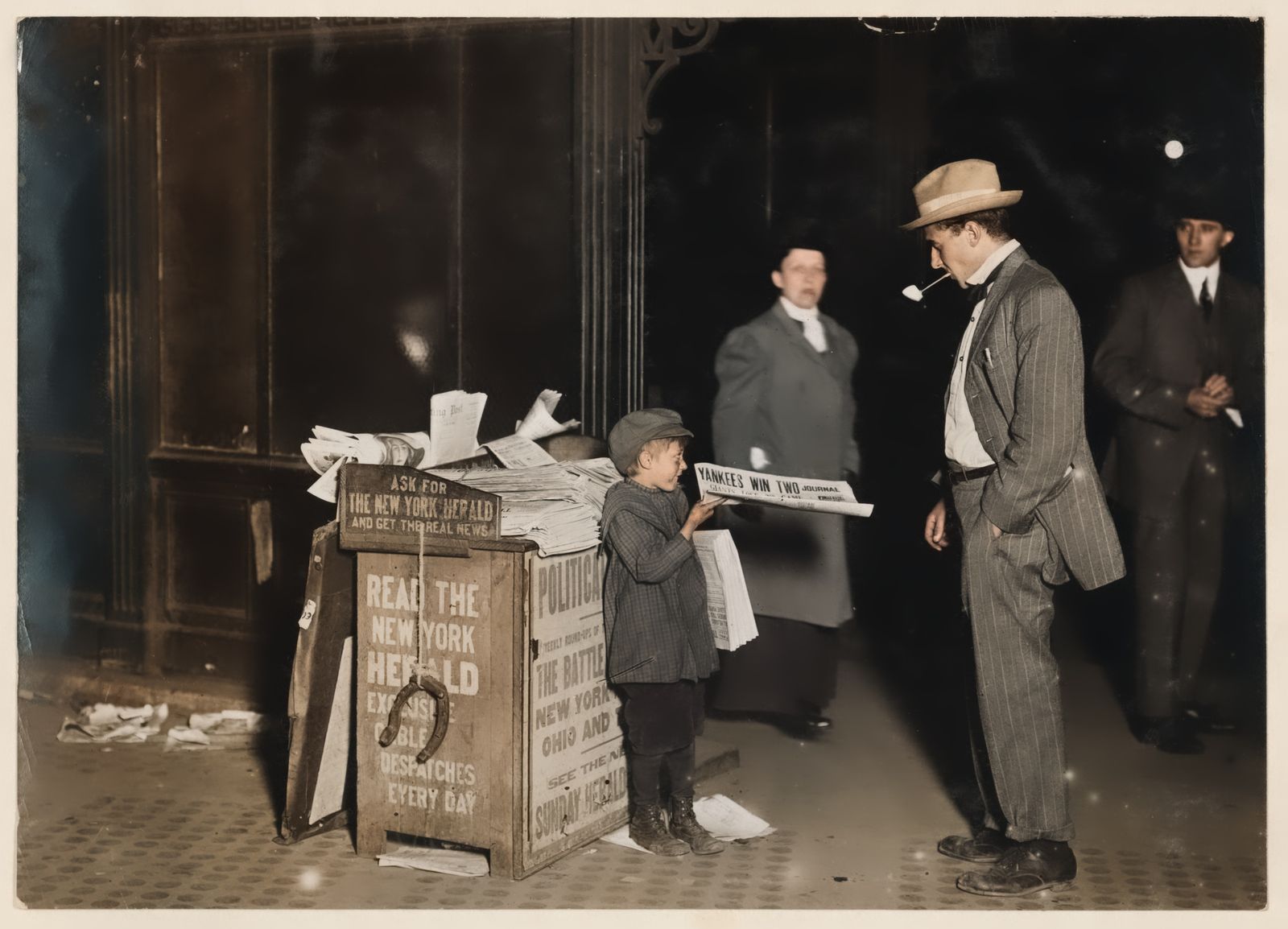 A sepia 1910 night street photograph of a newsboy at his stand with three adults nearby, before and after AI colourisation and contrast repair — after