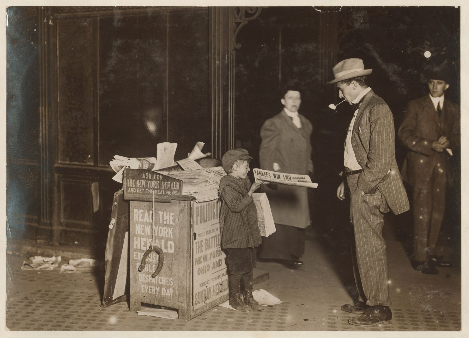 A sepia 1910 night street photograph of a newsboy at his stand with three adults nearby, before and after AI colourisation and contrast repair — before