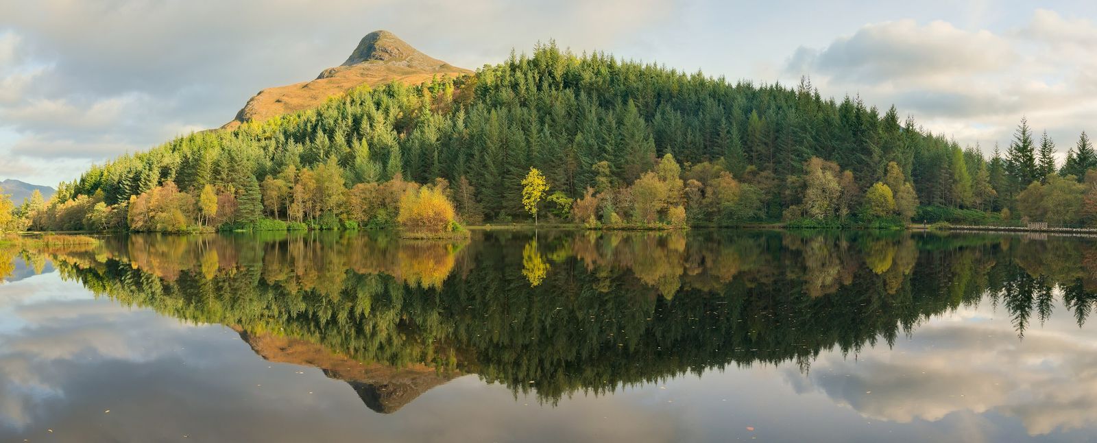 A Scottish loch with a wooded hill reflected in the water, before and after changing the season from summer green to autumn colours with AI — before